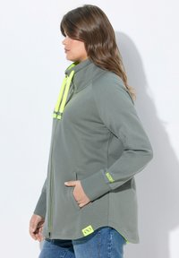 Chaqueta gris con cremallera y cuello alto, que presenta detalles en neón amarillo en la cremallera y la manga. Bolsillos laterales y un dobladillo curvado.