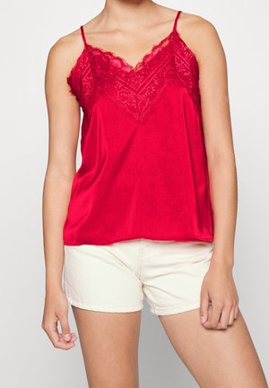 Kobieta w czerwonym jedwabnym podkoszulku z koronkową lamówką i beżowych, casualowych szortach na jednolitym jasnym tle.