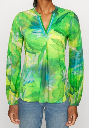 Vrouw draagt een lange mouwen groene blouse met een bladerenpatroon en blauwe bies bij de V-hals, gecombineerd met een blauwe spijkerbroek.