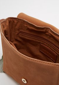 Ruskea mokkanahkainen käsilaukku, jossa on sileä sisus, varustettuna vetoketjullisella taskulla ja napsautettavalla sulkimella. Pintakuvioinnilla varustetut yksityiskohdat ulkopuolella lisäävät yksityiskohtia.