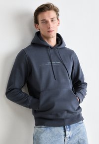 Donkergrijze hoodie met een voorzak en een capuchon met trekkoord. Beschikt over witte geborduurde logo-tekst en geribbelde mouwen. Draag met lichtblauwe jeans.
