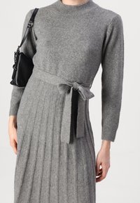 Vestito grigio in maglia con una texture a coste, scollatura rotonda, maniche lunghe e dettaglio della cintura in vita. Abbinato a una piccola borsa a spalla nera.