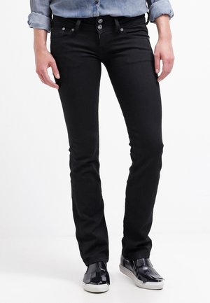 Henkilö pukeutunut mustiin slim-fit-farkkuihin, vaaleansiniseen pitkähihainen hihallinen käärittyyn paitaan ja mustiin slip-on-kenkiin, joissa on valkoiset pohjat.