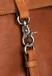 Bruine leren tas close-up met een metalen sluiting en riemverbinding, met een gestructureerd oppervlak en zichtbare stiksels.