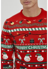 Roter Pullover mit festlichen Mustern, darunter Schneemänner, Weihnachtslichter, Zuckerstangen und der Text "FROHE WEIHNACHTEN" in grünen und weißen Akzenten.