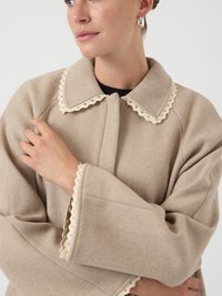 Beige vlněný kabát s širokým límcem zdobeným krajkovým lemem. Volný střih a široké rukávy s krajkovými detaily na manžetách. Hladká textura.