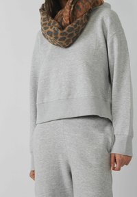 Personne portant un sweat-shirt et un pantalon gris clair avec une écharpe à imprimé léopard marron et noir, debout devant un fond uni.