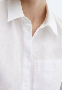 Camisa blanca de lino con cuello, que presenta un cierre frontal de botones, un bolsillo en el pecho y una tela suave con una ligera textura.