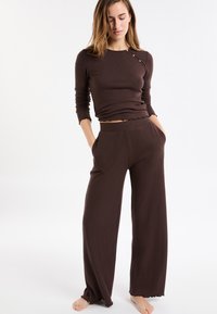 Braunes, geripptes Langarmoberteil mit gewelltem Saum und seitlichen Knopfdetails, kombiniert mit passenden, weit geschnittenen Hosen, beide aus weichem Stoff gefertigt.