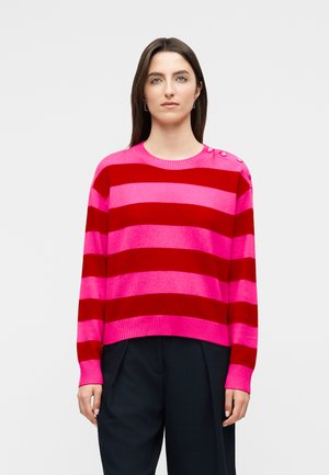 Femme aux cheveux lisses et foncés portant un pull large rayé rose et rouge avec des boutons sur l'épaule et un pantalon noir, debout devant un fond blanc.