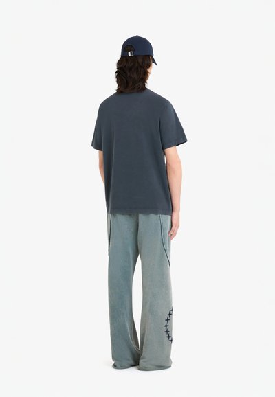 T-shirt à manches courtes gris foncé associé à un pantalon léger vert clair à coupe décontractée avec des poches latérales et un motif circulaire marine sur la jambe.