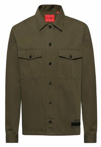 Camisa de algodón verde oliva de botones, con dos bolsillos en el pecho, botones negros y una etiqueta roja prominente con la marca 'HUGO'.