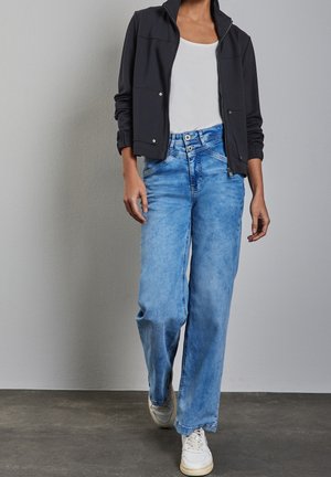 Personne marchant portant une veste zippée noire sur un haut blanc, un jean taille haute bleu clair à jambes larges, et des baskets blanches sur un sol gris.