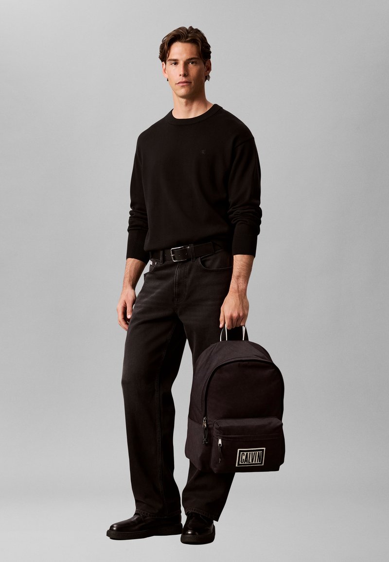 Jeune homme en tenue entièrement noire tenant un sac à dos Calvin noir par sa poignée sur un fond gris uni.