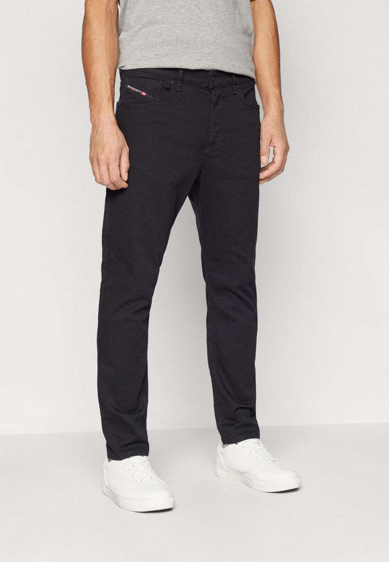 Jean noir en denim, coupe slim, avec cinq poches et une fermeture éclair. Porté avec des baskets blanches et un t-shirt gris.