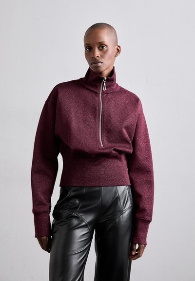 Bordowy, krótki polarowy sweatshirt z wysokim kołnierzem i zasuwakiem na pół, z długimi rękawami i miękkim, teksturowanym materiałem. W komplecie z czarnymi skórzanymi spodniami.