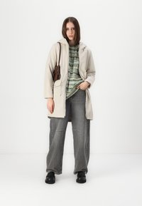 Lys beige hettejakke med lommer, båret over en grønnmønstret genser, kombinert med gråstripete bukser og svarte sko.