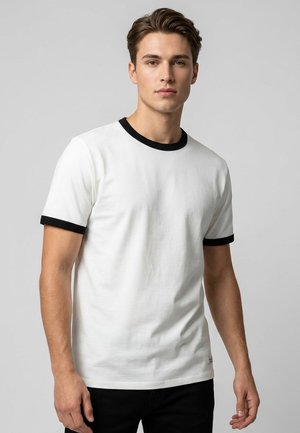 Junger Mann trägt ein weißes kurzärmeliges T-Shirt mit schwarzem Besatz am Kragen und an den Ärmeln, steht vor grauem Hintergrund.