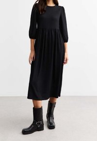 Mustapintainen ribbimäisen midi-mekko, jossa on kolmen neljänneksen hihat, vyötäröä korostava leikkaus ja levenevä helma, yhdistettynä mustiin nilkkabootseihin, joissa on metallikoristeita.