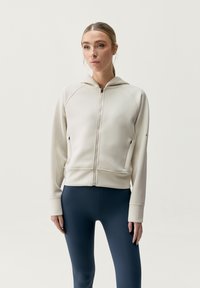 Felpa zip con cappuccio di un beige chiaro, con maniche raglan e tasche laterali, abbinata a leggings blu scuro. Tessuto morbido al tatto.