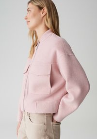 Veste bomber rose en tissu doux avec fermeture éclair, poignets côtelés, deux poches avant et coupe décontractée. Texture lisse et design minimaliste.