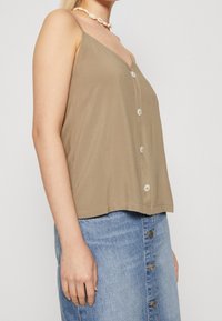 Žena v béžovém bezrukávovém topu s knoflíky a modré džíny sukni s knoflíky, doplněná bílým náhrdelníkem z mušlí.