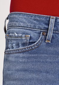 Ljust blå denimjeans med en framficka som har slitna detaljer och kontrasterande gula sömmar, visade mot en röd bakgrund.