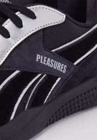 Čierne semišové tenisky so striebornými akcentmi, gumovou podrážkou a bielymi detailmi. Na boku sa nachádza značka „PLEASURES“.