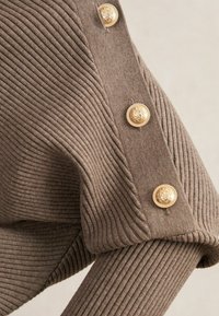 Ruskea ribbi-neulosmateriaali, jossa on kolme koristeellista kultanappia, joissa on vaakunamalli, vaatekappaleen hihassa.