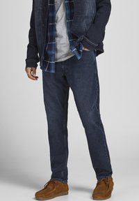 Vaqueros de mezclilla azul oscuro con un corte relajado, combinados con una camisa de cuadros bajo una chaqueta de mezclilla oscura y zapatos de ante marrones.