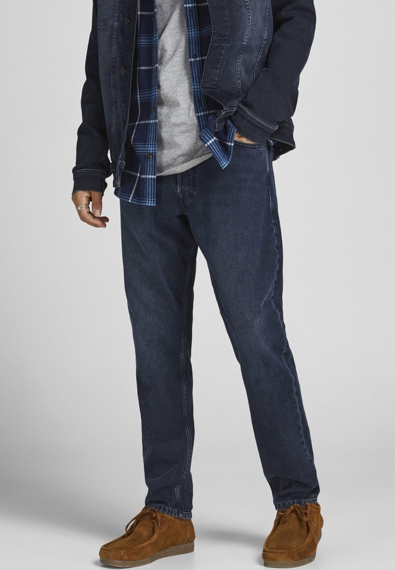 Vaqueros de mezclilla azul oscuro con un corte relajado, combinados con una camisa de cuadros bajo una chaqueta de mezclilla oscura y zapatos de ante marrones.