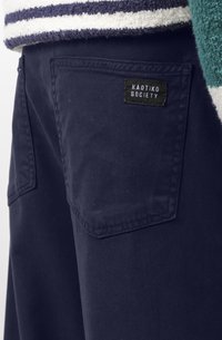 Pantalons bleu marine foncé avec une poche arrière portant une étiquette noire indiquant "KAOTIKO SOCIETY". Le matériau semble robuste avec une texture lisse.