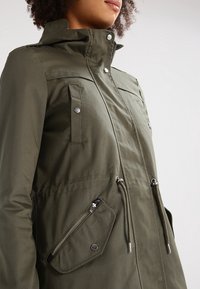 Veste vert olive avec un col montant, dotée de poches poitrine et latérales, d'accessoires argentés et de cordons ajustables à la taille.