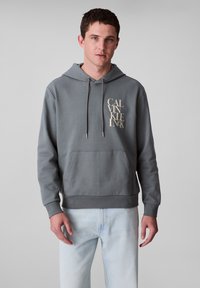 Felpa grigia con tasca anteriore e cappuccio regolabile con cordino. Presenta un grande logo beige di Calvin Klein sul lato sinistro del petto. Realizzata in materiale morbido.