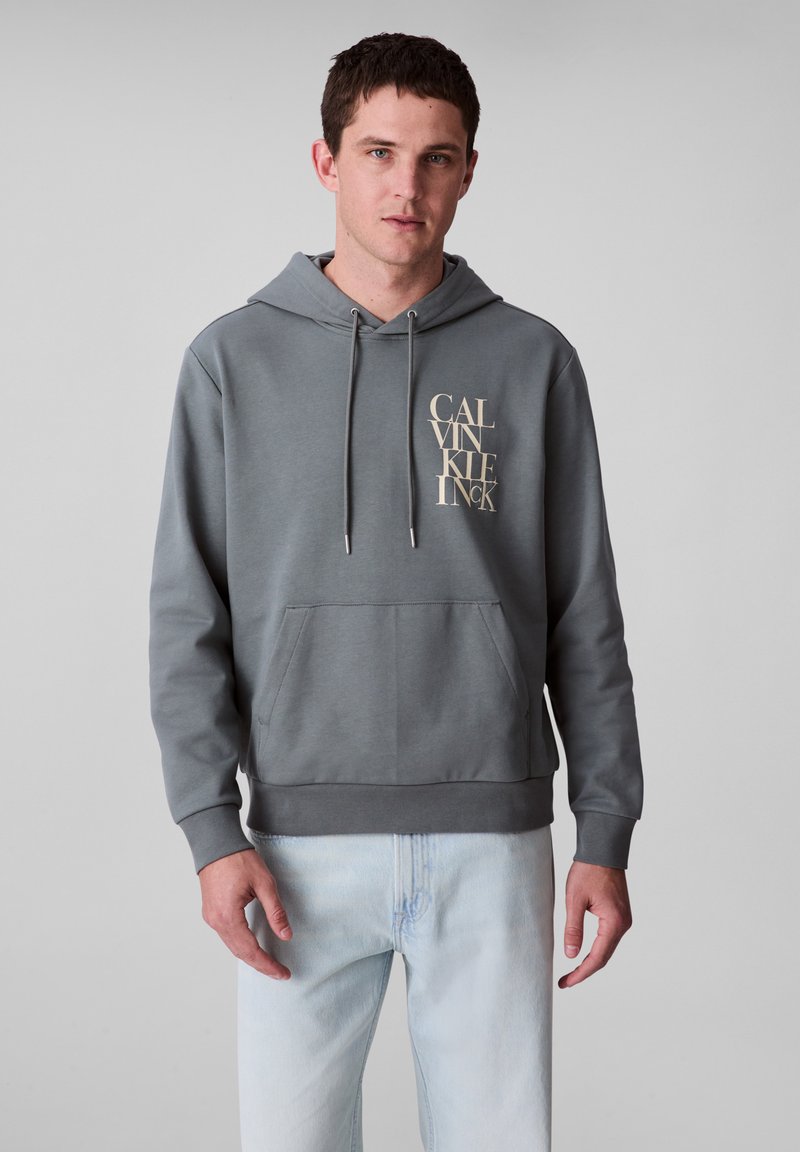 Felpa grigia con tasca anteriore e cappuccio regolabile con cordino. Presenta un grande logo beige di Calvin Klein sul lato sinistro del petto. Realizzata in materiale morbido.