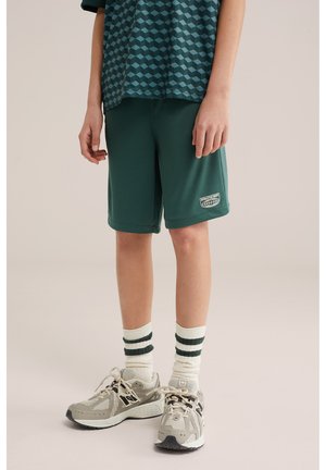 Niño con pantalones cortos deportivos verdes, camiseta verde oscuro con estampado, calcetines blancos con rayas verdes y zapatillas deportivas beige, de pie sobre un fondo liso.