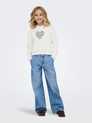 Ragazza sorridente con capelli biondi ricci che indossa una felpa bianca con un cuore a stampa leopardata e jeans blu a gamba larga con scarpe marroni.