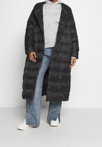 Abrigo acolchado negro con un corte largo y holgado, que cuenta con un cierre de botones a presión y un patrón de acolchado. Combinado con sudadera gris y jeans de pierna ancha.