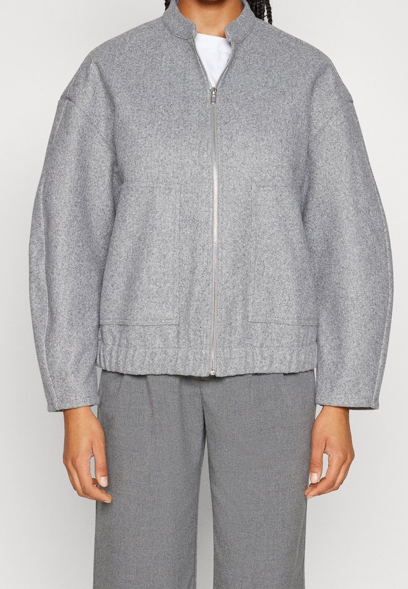 Personne portant une veste zippée gris clair à manches longues et un pantalon gris assorti, debout les bras détendus le long du corps.