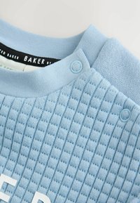 Lysblå tekstureret sweatshirt med ribbet krave og trykknapper. Har et quiltet mønster på kroppen og et sort mærke indeni.