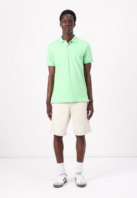Light green cotton polo shirt with short sleeves, paired with beige shorts. Features a simple design and worn with grey and white sneakers.