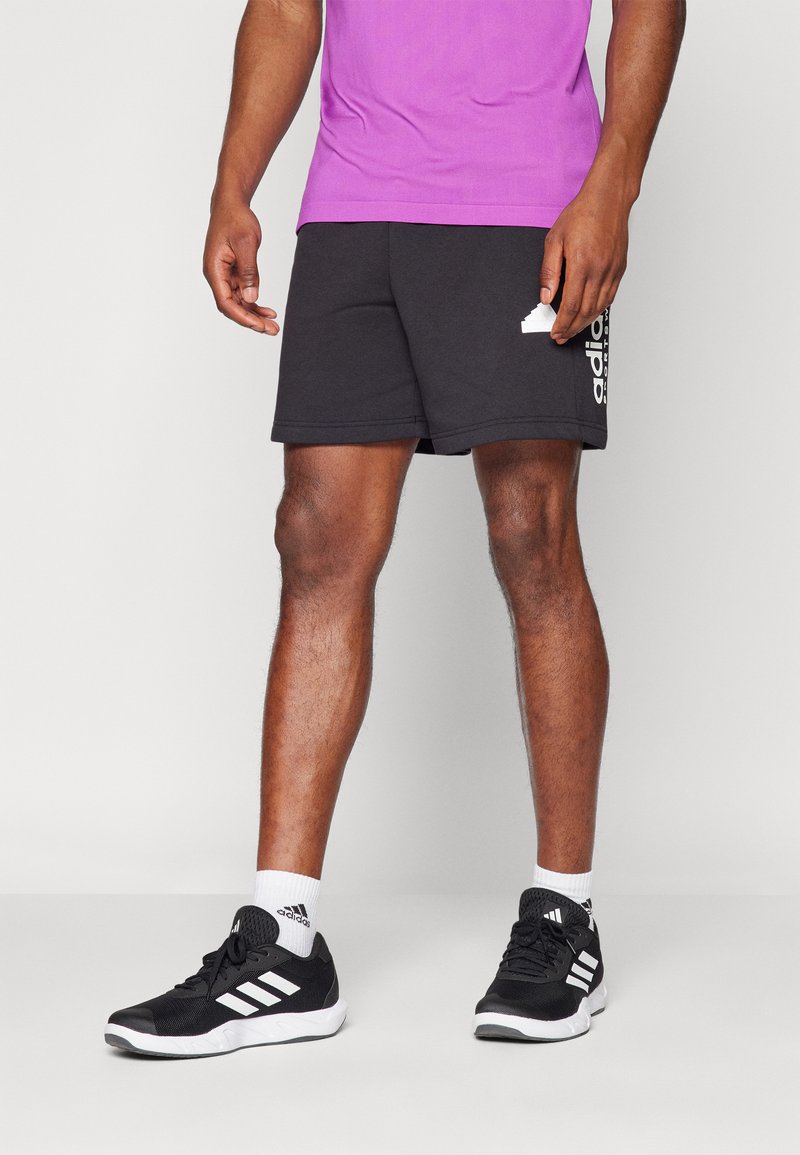Pantalones cortos deportivos negros con el logo blanco de Adidas, combinados con zapatillas deportivas negras que tienen tres rayas blancas y calcetines blancos tipo crew.