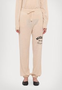 Bež trenerke z vrvico v pasu, s pripetimi gležnji in črnim vezenim logotipom "MOSCHINO Jeans" na strani. Mehka tekstura tkanine.