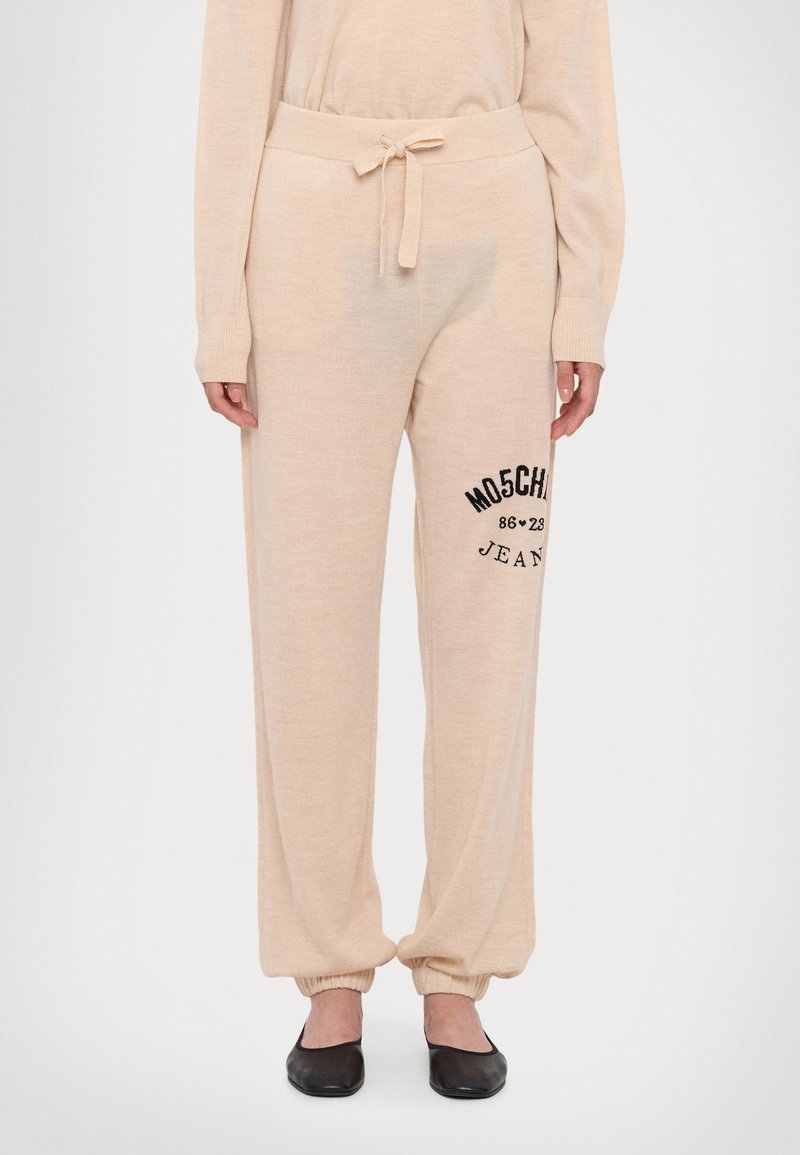 Bež trenerke z vrvico v pasu, s pripetimi gležnji in črnim vezenim logotipom "MOSCHINO Jeans" na strani. Mehka tekstura tkanine.