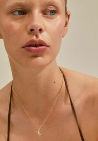 Collier en or avec un pendentif en forme de croissant de lune, porté sur une peau nue avec une lanière marron, présentant une texture lisse et un design délicat.