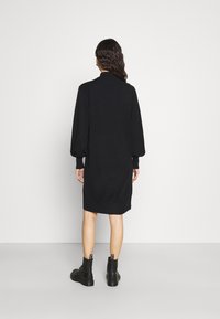 Robe noire oversized avec de longues manches bouffantes et un ourlet côtelé. Le modèle porte des bottes de combat noires, mettant en avant un design décontracté et ample.