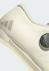 Hellbeige Sportschuh mit einer strukturierten Oberseite, runder Form, dezentem Logo und einem runden Drehknopf zur Anpassung der Passform. Glattes Sohlen-Detail.