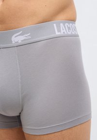 Gros plan d'une personne portant un boxer Lacoste gris clair avec une ceinture de marque et le logo crocodile.