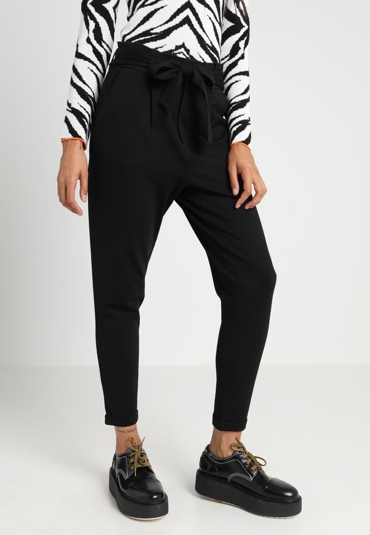Zwarte tapered broek van zacht materiaal, met een gestrikt taille, zijzakken en een soepele textuur. Gecombineerd met glanzende zwarte plateauschoenen.