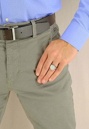 Pantaloni di cotone grigi con dettagli di cucitura visibili, una cintura di pelle nera con una fibbia metallica e un anello d'argento con una superficie rotonda e testurizzata.